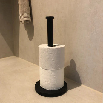 Un porte-rouleau de papier toilette Jurado Black de yessential.fr se dresse verticalement dans le coin d'une salle de bain carrelée beige, contenant deux rouleaux de papier toilette blanc, avec une serviette soigneusement pliée sur le mur au-dessus.