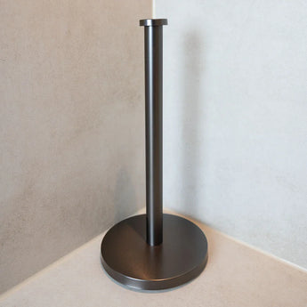Le porte-serviettes en papier Jurado Gunmetal de yessential.fr, avec une base ronde et une tige verticale, repose sur un comptoir léger et complète d'autres pièces Jurado pour un look cohérent contre les murs beige clair.