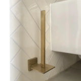 Le porte-rouleau papier toilette Bellino Gold de yessential.fr, au design moderne et finition dorée, possède une tige verticale et une base carrée. Il se monte au mur sur des carreaux diagonaux blancs près de toilettes blanches et ajoute un style minimal à votre espace.