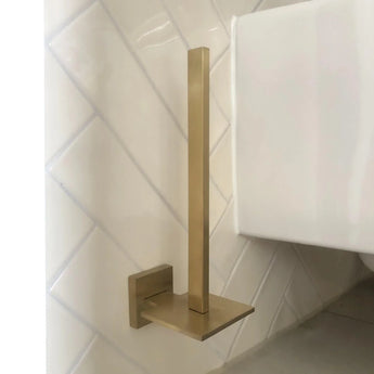 Le porte-rouleau papier toilette Bellino Gold de yessential.fr, au design moderne et finition dorée, possède une tige verticale et une base carrée. Il se monte au mur sur des carreaux diagonaux blancs près de toilettes blanches et ajoute un style minimal à votre espace.