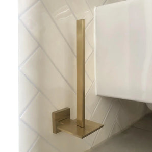Le porte-rouleau papier toilette Bellino Gold de yessential.fr, au design moderne et finition dorée, possède une tige verticale et une base carrée. Il se monte au mur sur des carreaux diagonaux blancs près de toilettes blanches et ajoute un style minimal à votre espace.