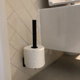 Un rouleau de papier toilette est suspendu verticalement au porte-rouleau de rechange Bellino Black de yessential.fr, fixé au mur carrelé d'une salle de bains à côté de toilettes blanches.