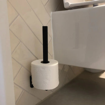 Un rouleau de papier toilette est suspendu verticalement au porte-rouleau de rechange Bellino Black de yessential.fr, fixé au mur carrelé d'une salle de bains à côté de toilettes blanches.