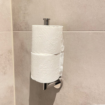 Deux rouleaux de papier toilette empilés sur le porte-rouleau de papier toilette Roberto Chrome de yessential.fr, monté contre un mur de salle de bains carrelé beige clair.