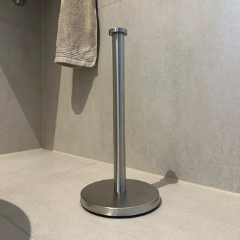 Le porte-rouleau de papier toilette autoportant en acier inoxydable Jurado yessential.fr a une construction en acier inoxydable avec une base ronde et une tige verticale, placée sur un sol carrelé de salle de bain. Une serviette beige est accrochée au mur en arrière-plan.