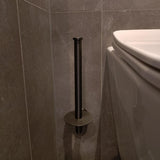 Le porte-rouleau de papier toilette vertical Roberto Gunmetal yessential.fr est monté sur un mur carrelé gris à côté de toilettes blanches. Le conteneur vide projette une ombre sur le mur et le sol.
