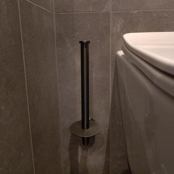 Le porte-rouleau de papier toilette vertical Roberto Gunmetal yessential.fr est monté sur un mur carrelé gris à côté de toilettes blanches. Le conteneur vide projette une ombre sur le mur et le sol.