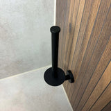 Le porte-rouleau de rechange yessential.fr Roberto Black est monté verticalement sur un mur de salle de bain à motif bois, photographié d'en haut avec des murs et un sol carrelés en arrière-plan.