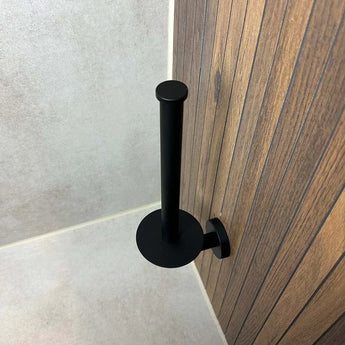Le porte-rouleau de rechange yessential.fr Roberto Black est monté verticalement sur un mur de salle de bain à motif bois, photographié d'en haut avec des murs et un sol carrelés en arrière-plan.