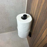 Le porte-rouleau de rechange Roberto Black de yessential.fr est monté verticalement sur le mur de la salle de bain avec carrelage et boiseries beiges et un flux continu de papier toilette.