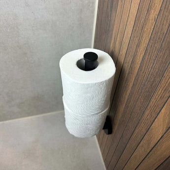 Le porte-rouleau de rechange Roberto Black de yessential.fr est monté verticalement sur le mur de la salle de bain avec carrelage et boiseries beiges et un flux continu de papier toilette.