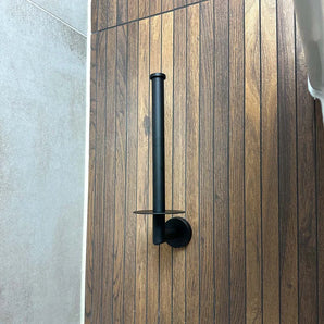 Le porte-rouleau de rechange Roberto Black de yessential.fr est monté verticalement sur un mur avec des panneaux imitation bois, à côté de carrelage gris clair.