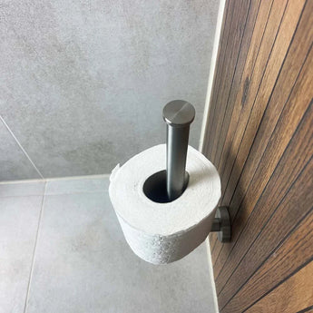Le porte-rouleau papier toilette Roberto en acier inoxydable de yessential.fr avec revêtement noir mat est monté verticalement sur un mur en bois dans une salle de bain aux murs et au sol carrelés gris clair.