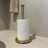 L'ensemble WC Ludo Gold (sans perçage) de yessential.fr se dresse sur un sol de salle de bain légèrement carrelé avec deux rouleaux de papier toilette et une serviette beige sur le mur en arrière-plan - le tout installable sans perçage.