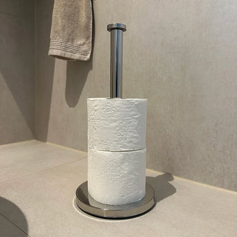 L'ensemble de toilettes Delo Chrome (sans perçage) de yessential.fr se dresse sur un sol de salle de bain légèrement carrelé avec deux rouleaux de papier toilette en relief, tandis qu'une serviette beige est suspendue en arrière-plan.