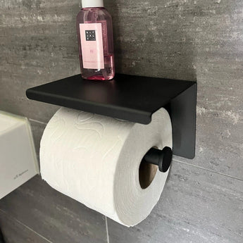 Le porte-rouleau de papier toilette Tazio Black (No Drill) de yessential.fr, avec un rouleau de papier toilette blanc et une bouteille rose, repose contre un mur de salle de bain carrelé gris.