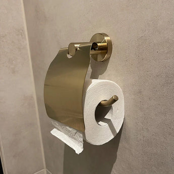 L'ensemble WC Lauro Gold de yessential.fr est parsemé d'un porte-rouleau papier toilette avec couvercle, monté sur un mur de couleur beige. Le processus d'installation est terminé, avec un papier de tri selon le processus d'installation.