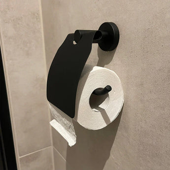 Un rouleau de papier toilette pend de manière inégale à un support pour set de toilette Sandro Black de yessential.fr, monté latéralement dans la salle de bain de manière à ce que le support recouvre partiellement le rouleau.