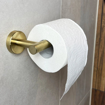 Un rouleau de papier toilette blanc gaufré est suspendu au porte-rouleau de papier toilette Tino Gold de yessential.fr contre un mur carrelé marron clair, avec une feuille volante ajoutant une touche de style accessoire de toilette de luxe à l'espace.