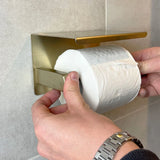 Une personne portant une montre en argent place du papier toilette sur l'ensemble de toilettes Cano Gold (sans perçage) de yessential.fr, monté sur un mur carrelé.