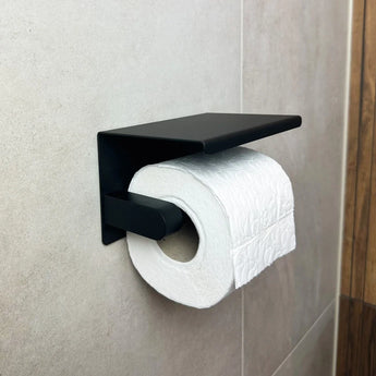 Un rouleau de papier toilette blanc sur l'ensemble Armando Black de yessential.fr - et supportez l'ensemble de toilette noir mat élégant avec une étagère rectangulaire, élégamment monté sur mur carrelé pour une finition élégante à vos accessoires de toilette noirs.