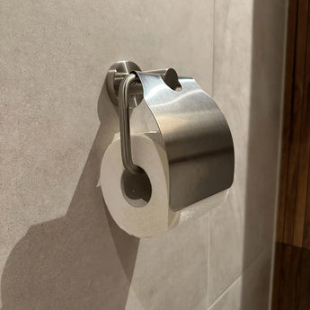 Un ensemble de WC Lauro et un en acier inoxydable de yessential.fr est monté sur le mur de la baignoire carrelé beige avec les panneaux et bois apparents, avec un porte-papier toilette avec couvercle et métal brossé - partie d'une élégante collection d'accessoires de baignoire.