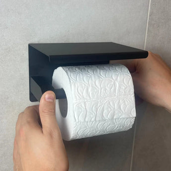 Les mains d'une personne placent un rouleau de papier toilette blanc gaufré sur l'ensemble de toilette Juano Black (sans perçage) de yessential.fr, qui se monte sur un mur carrelé de couleur claire sans perçage.