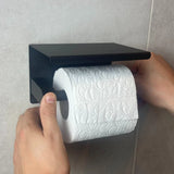 Deux mains placent un rouleau de papier toilette à motifs sur l'ensemble de toilette noir Delo (sans perçage) de yessential.fr, monté contre un mur carrelé - un ensemble d'accessoires de toilette noir élégant et facile à installer sans perçage.