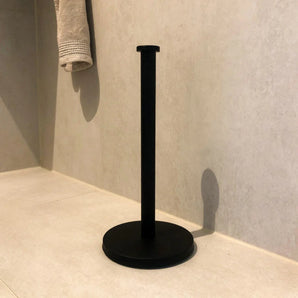 Le Jurado Black de yessential.fr, un porte-rouleau de papier toilette autoportant minimaliste, se dresse sur le sol de la salle de bain de couleur claire, contre le mur, avec une serviette beige au-dessus à gauche.