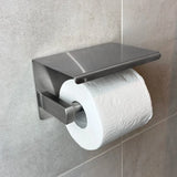 Un rouleau de papier toilette sur un porte-rouleau de papier toilette en acier inoxydable Tazio yessential.fr avec étagère, monté sur un mur de salle de bains carrelé gris clair.