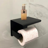 Le porte-rouleau papier toilette Tazio Black de yessential.fr se fixe sur un mur carrelé, avec un rouleau de papier toilette blanc à coeurs roses et une pompe à savon ou lotion sur l'étagère.