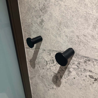 Quatre crochets à serviettes Carreno Black de yessential.fr sont montés sur un mur en pierre gris clair texturé avec des ombres projetées par une lumière oblique. Sur la gauche, un cadre métallique vertical est visible – un accessoire idéal pour la salle de bain.