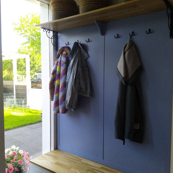 Un couloir avec un banc en bois, un mur bleu clair avec la patère Pagano Black de yessential.fr contenant trois manteaux et une étagère au-dessus. La lumière du soleil traverse une porte ouverte, révélant un jardin verdoyant et des fleurs à l’extérieur.