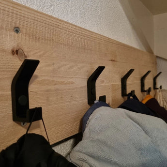 Une rangée de 10 crochets Huso Black de yessential.fr est montée sur une étagère en bois clair et maintient les manteaux et les vestes contre un mur texturé blanc pour un look épuré et moderne.