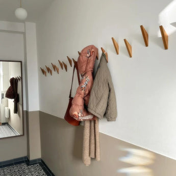 Une entrée avec le portemanteau minimaliste Fabio Oak de yessential.fr avec des crochets géométriques sur un mur blanc. Un manteau rose, une cape matelassée beige, une écharpe marron et un sac marron y sont suspendus. Le miroir et le carrelage sont visibles.
