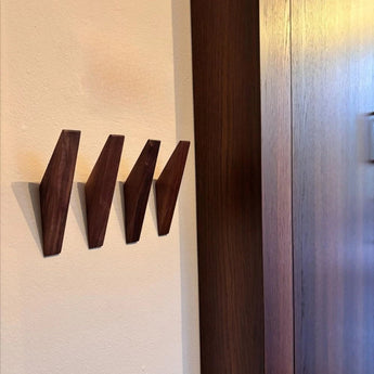 Trois patères modernes Fabio Walnut de yessential.fr sont montées sur un mur beige à côté du bois sombre, projetant des ombres douces et créant un look minimaliste de porte-manteau en bois.