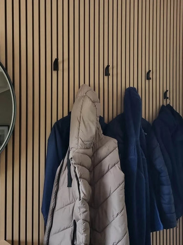 Un mur en lattes de bois est équipé de quatre crochets yessential.fr Huso Black, chacun retenant un manteau ou une veste beige ou bleu. Un miroir rond est accroché sur le côté gauche du mur.
