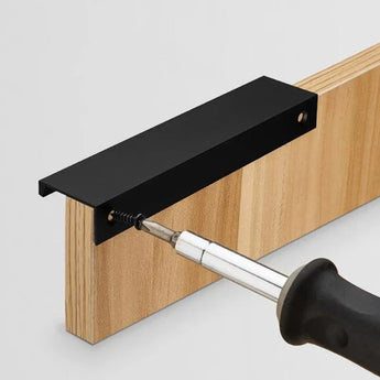 Gros plan d'une main utilisant un tournevis pour enfoncer une vis à travers un support métallique Enzo Black de yessential.fr jusqu'au bord d'une planche de bois. Le support est conçu à des fins de montage ou de connexion.