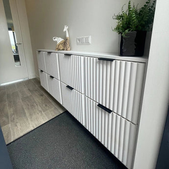 Dans un hall moderne se trouve le meuble nervuré Enzo Black de yessential.fr avec des poignées d'angle noires, représentant une fougère en pot et une sculpture de girafe. Le parquet clair se transforme en moquette sombre près du placard.