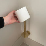 Une main place un rouleau de papier toilette sur le porte-rouleau de papier toilette Roberto Gold de yessential.fr, un support incliné de couleur dorée qui se fixe au mur de la salle de bain - idéal pour améliorer vos accessoires de salle de bain.