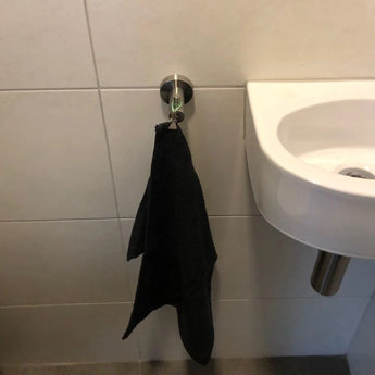 Une serviette noire est suspendue à un crochet Justo en acier inoxydable de yessential.fr sur un mur carrelé de salle de bains à côté d'un petit lavabo blanc avec un tuyau d'évacuation en métal.
