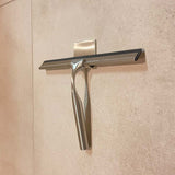 Le Zamoro Stainless Steel de yessential.fr, un extracteur de douche en acier inoxydable avec lame en caoutchouc noir, s'accroche à un crochet mural avec un système d'accrochage 3M sur du carrelage beige.