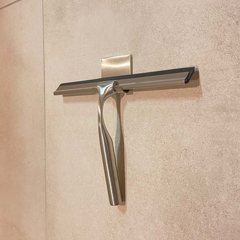 Le Zamoro Stainless Steel de yessential.fr, un extracteur de douche en acier inoxydable avec lame en caoutchouc noir, s'accroche à un crochet mural avec un système d'accrochage 3M sur du carrelage beige.
