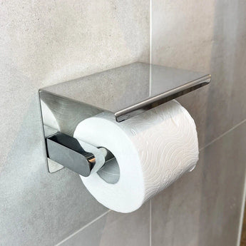 Un rouleau de papier toilette se trouve sur l'ensemble WC Ludo Chrome (sans perçage) de yessential.fr - un support moderne en acier inoxydable avec un dessus plat, fixé à un mur carrelé gris sans perçage.