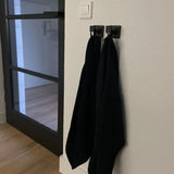 Deux serviettes noires sont suspendues côte à côte sur des crochets noirs Valo de yessential.fr dans un intérieur minimaliste, les crochets étant montés sur un mur blanc près d'une porte vitrée moderne.