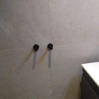 Deux crochets Justo Black Hook de yessential.fr sont montés sur un mur carrelé beige, projetant des ombres à paupières. Un comptoir blanc et un meuble qui s'intègre parfaitement.