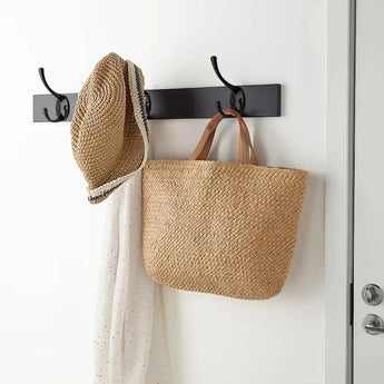 Le Nismo Black 1-Hook de yessential.fr est fixé au mur et contient un chapeau de paille, une écharpe blanche à petits points noirs et un sac fourre-tout en paille tressée avec des poignées marron contre un mur blanc.