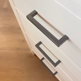 Gros plan de trois tiroirs blancs avec des poignées modernes Vasco Gunmetal de yessential.fr, sur un parquet en bois clair. Les tiroirs ont un léger angle, ce qui fait ressortir les lignes épurées et le design minimaliste.