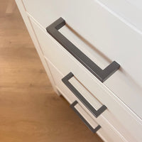 Gros plan de trois tiroirs blancs avec des poignées modernes Vasco Gunmetal de yessential.fr, sur un parquet en bois clair. Les tiroirs ont un léger angle, ce qui fait ressortir les lignes épurées et le design minimaliste.