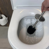 Une main équipée de la Brosse Extra yessential.fr, équipée d'une tête interchangeable, nettoie l'intérieur d'une cuvette de toilette blanche sous l'eau courante ; un rouleau de papier toilette est visible sur un support à proximité.